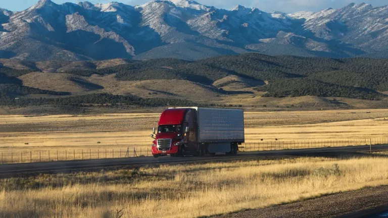 Caminhão vermelho com baú rodando em rodovia aberta, com montanhas ao fundo