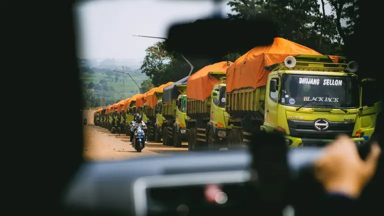 Fila de caminhões com lona laranja parados na beira de uma estrada de terra, vista de dentro de uma cabine